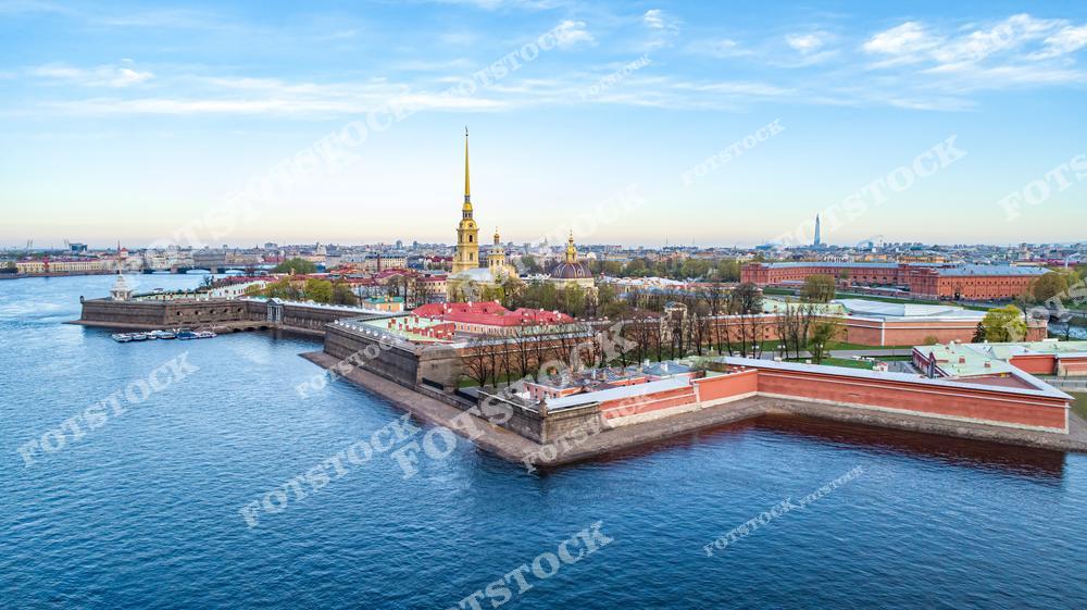 На снимке изображена Петропавловская крепость — одно из старейших архитектурных сооружений Санкт-Петербурга. Центральное место занимает золотой шпиль Петропавловского собора, возвышающегося над крепостными стенами. Река Нева окружает крепость, подчеркивая её историческую значимость и красоту. Весенний пейзаж с голубым небом добавляет легкости и света в общую композицию. Эта крепость — символ основания города и его неразрывной связи с историей.